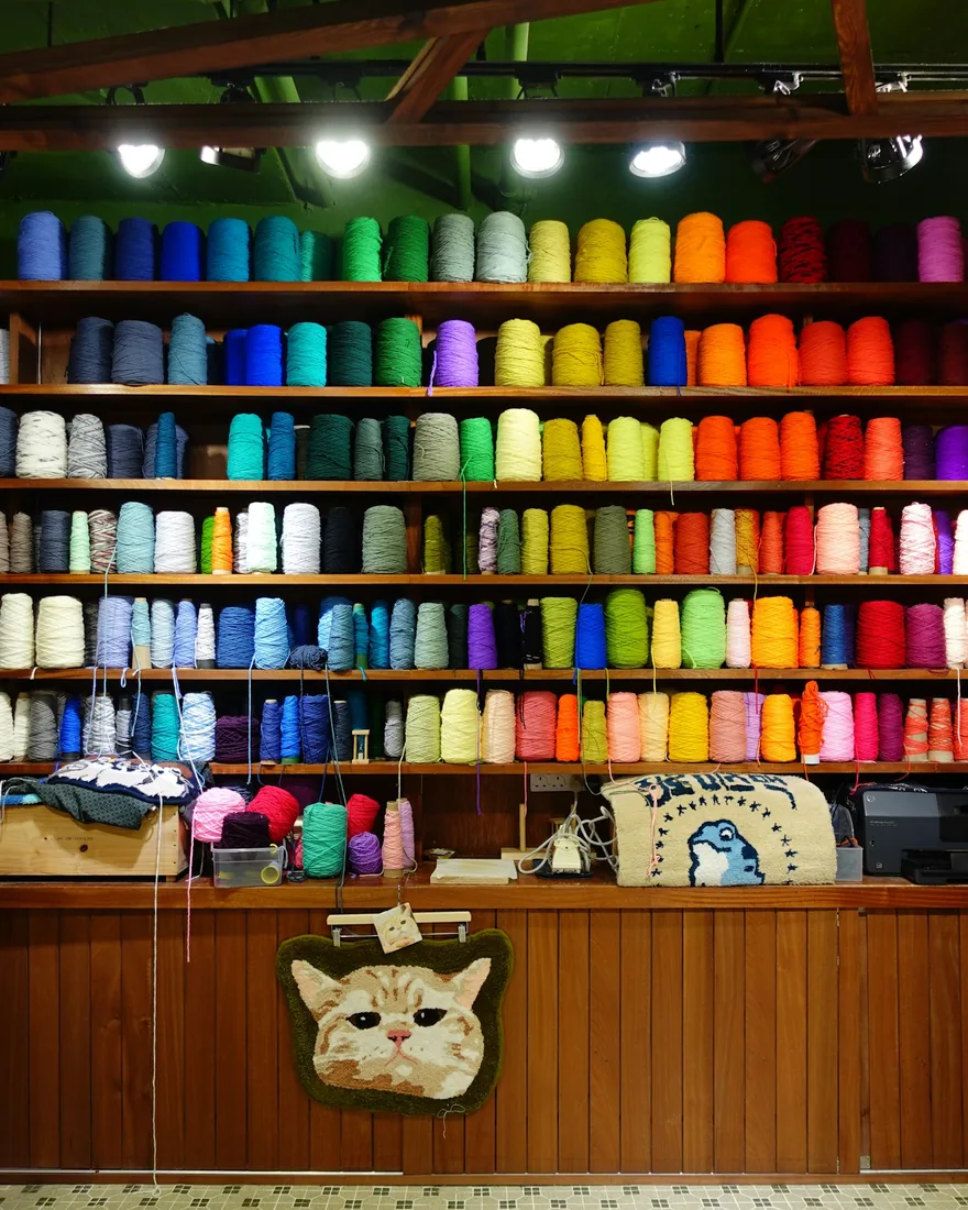 Estanterías de un taller de tufting con conos de hilo ordenados por color y piezas tuftadas colgadas debajo